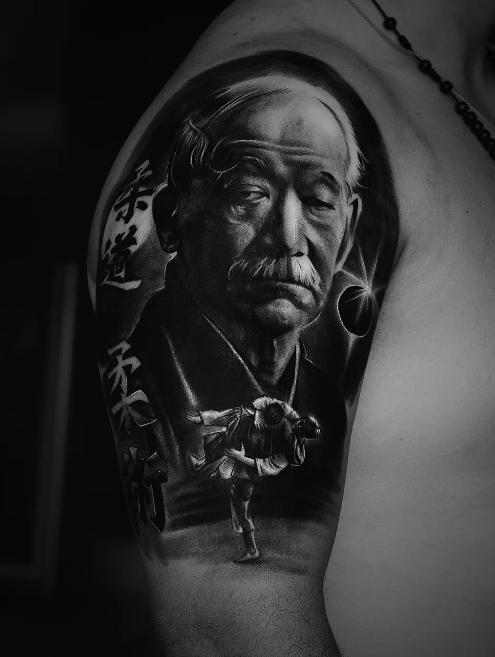Retrato realista de um mestre de artes marciais em preto e cinza, tributo tatuado no braço por Felipe Carlos.
