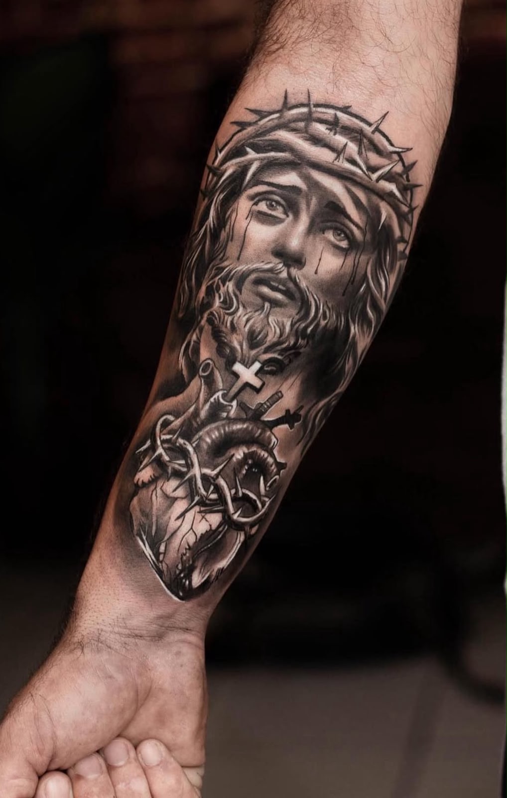 Antebraço com a face de Cristo coroado de espinhos sobre coração anatômico e cruz, realismo em preto e cinza por Felipe Carlos.